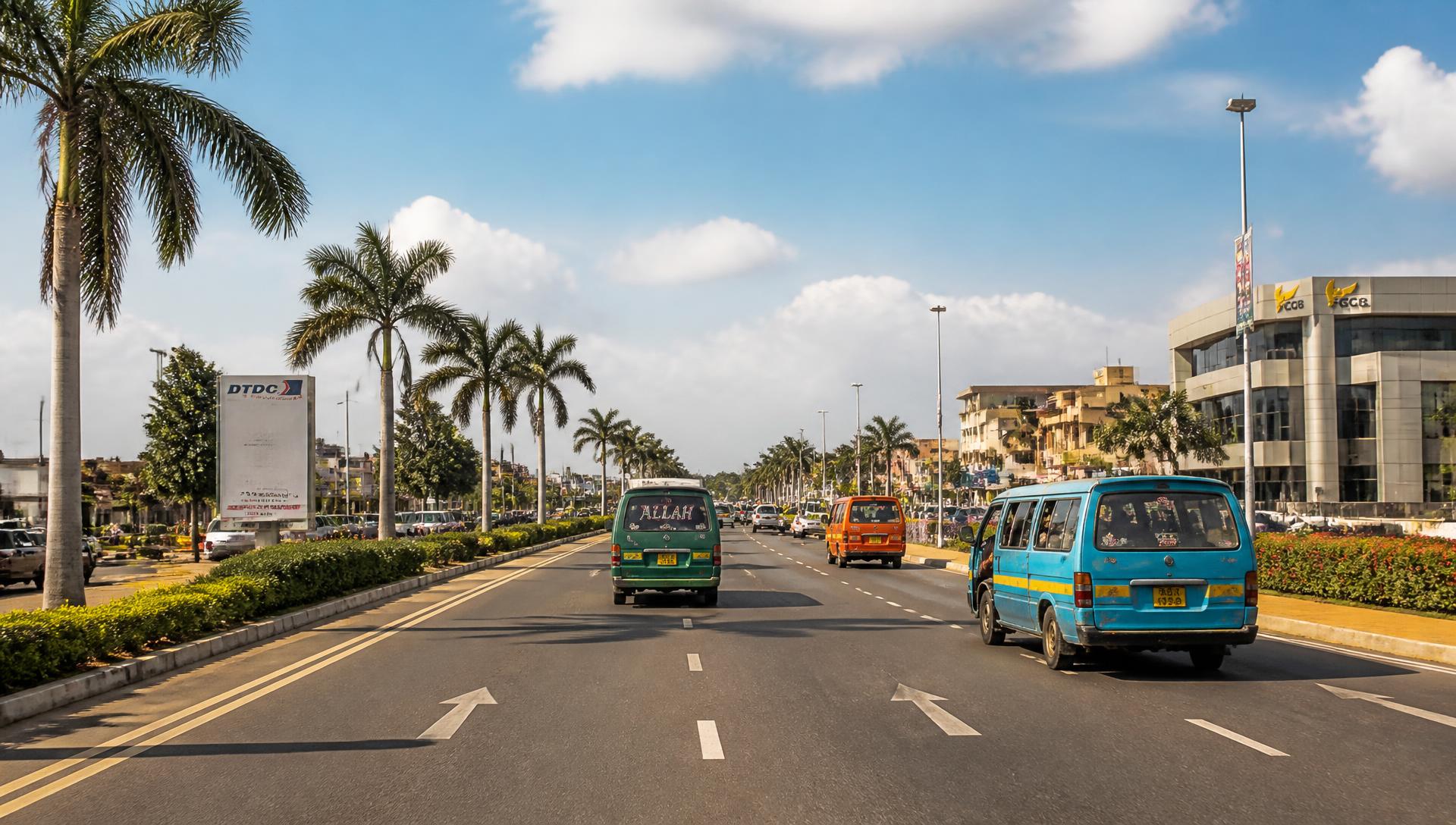 A palm-lined road in Tema, Ghana with minibuses and taxis under a bright blue sky
